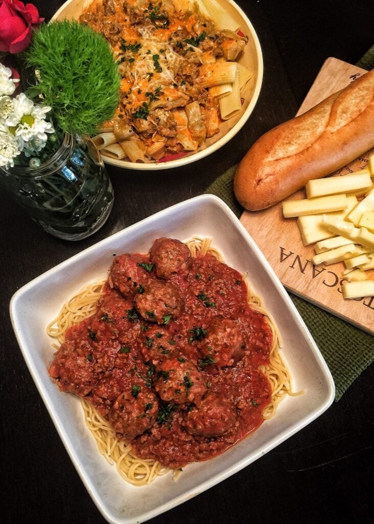 Two plates of pasta with different sauces next to a cutting board with a loaf of bread and pieces of cheese.