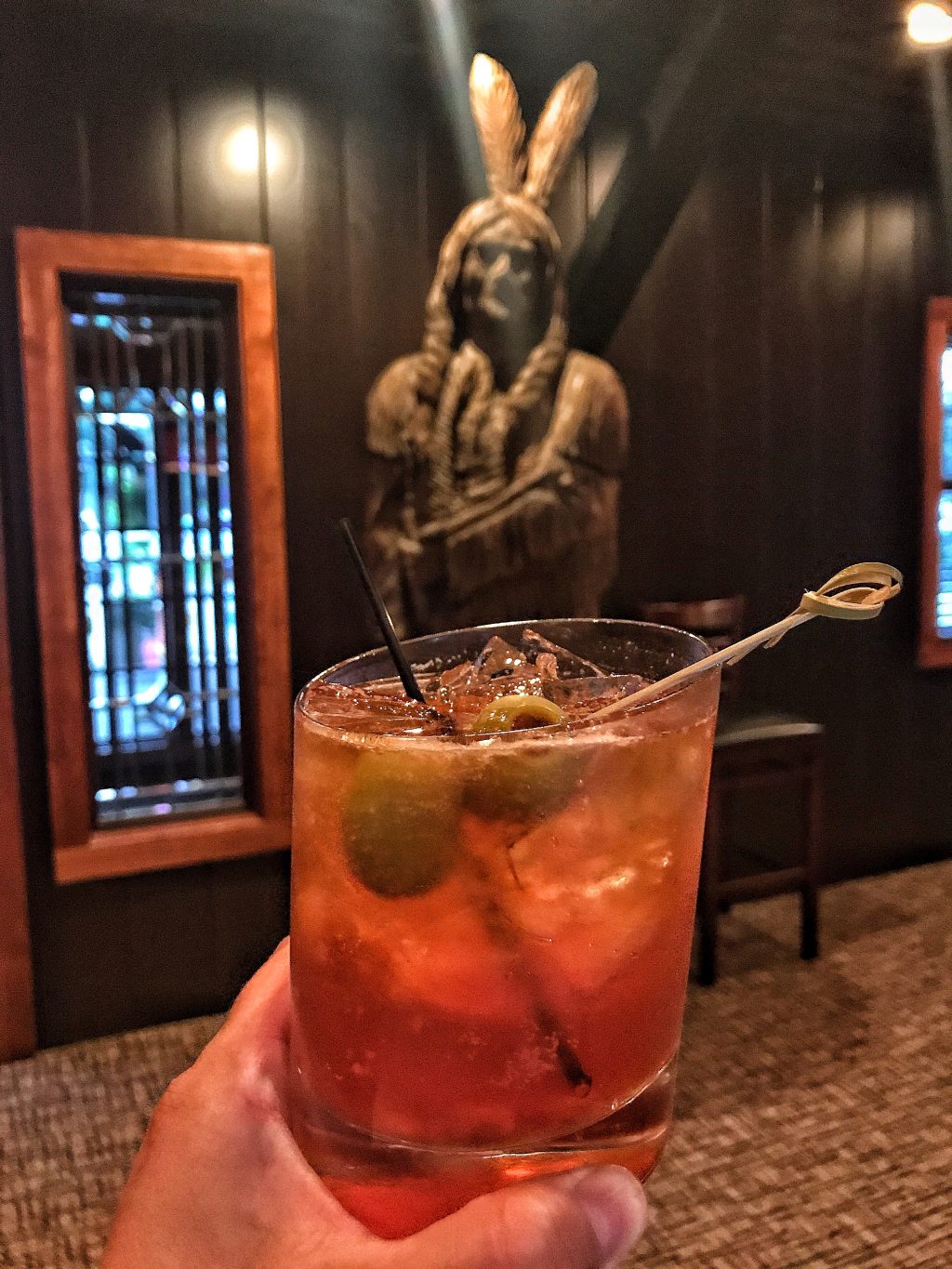 A brandy Old Fashioned being held up with a Native American chief statue in the background.
