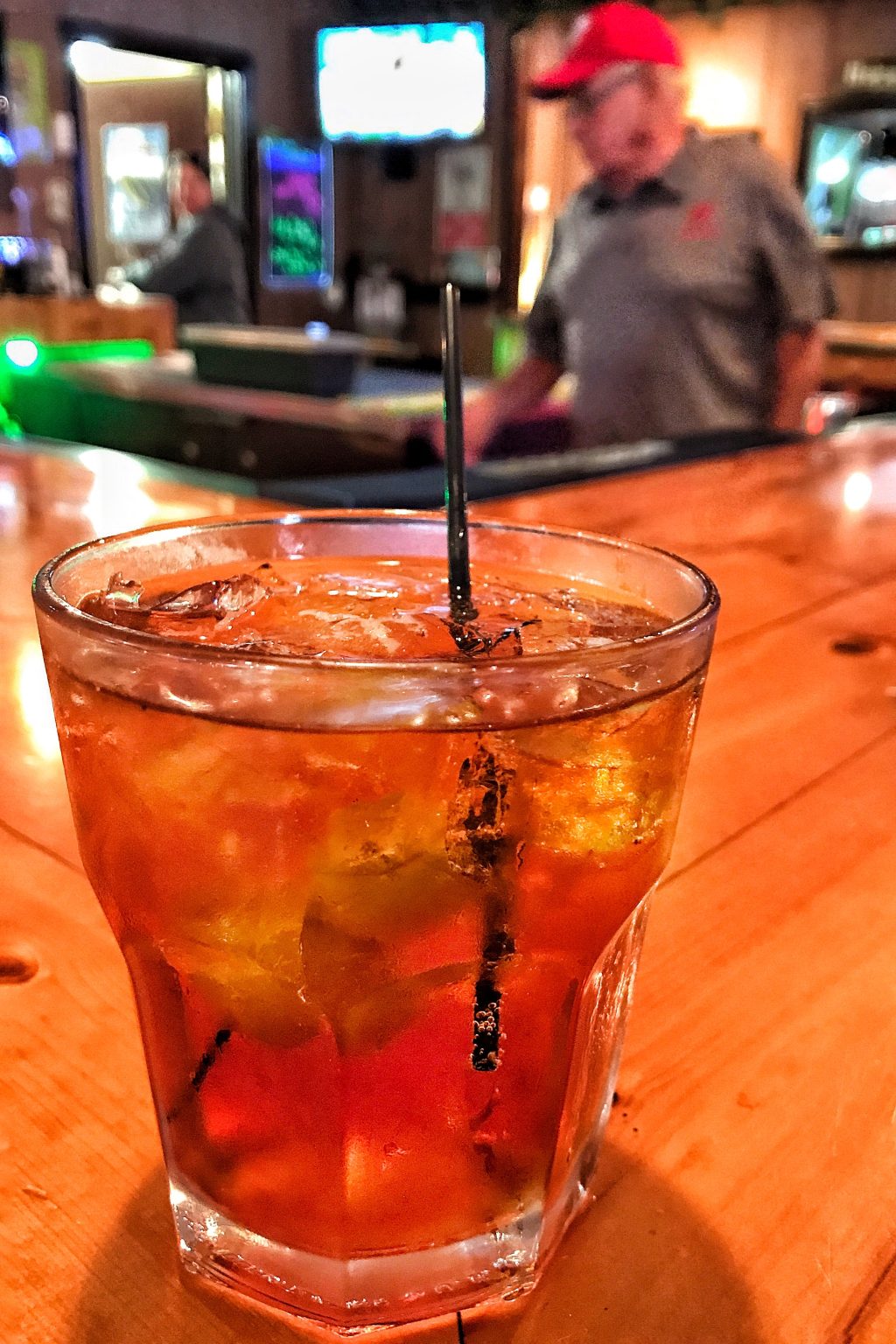 A brandy Old Fashioned on a bar with a bartender wearing a red baseball cap and glasses blurry in the background.
