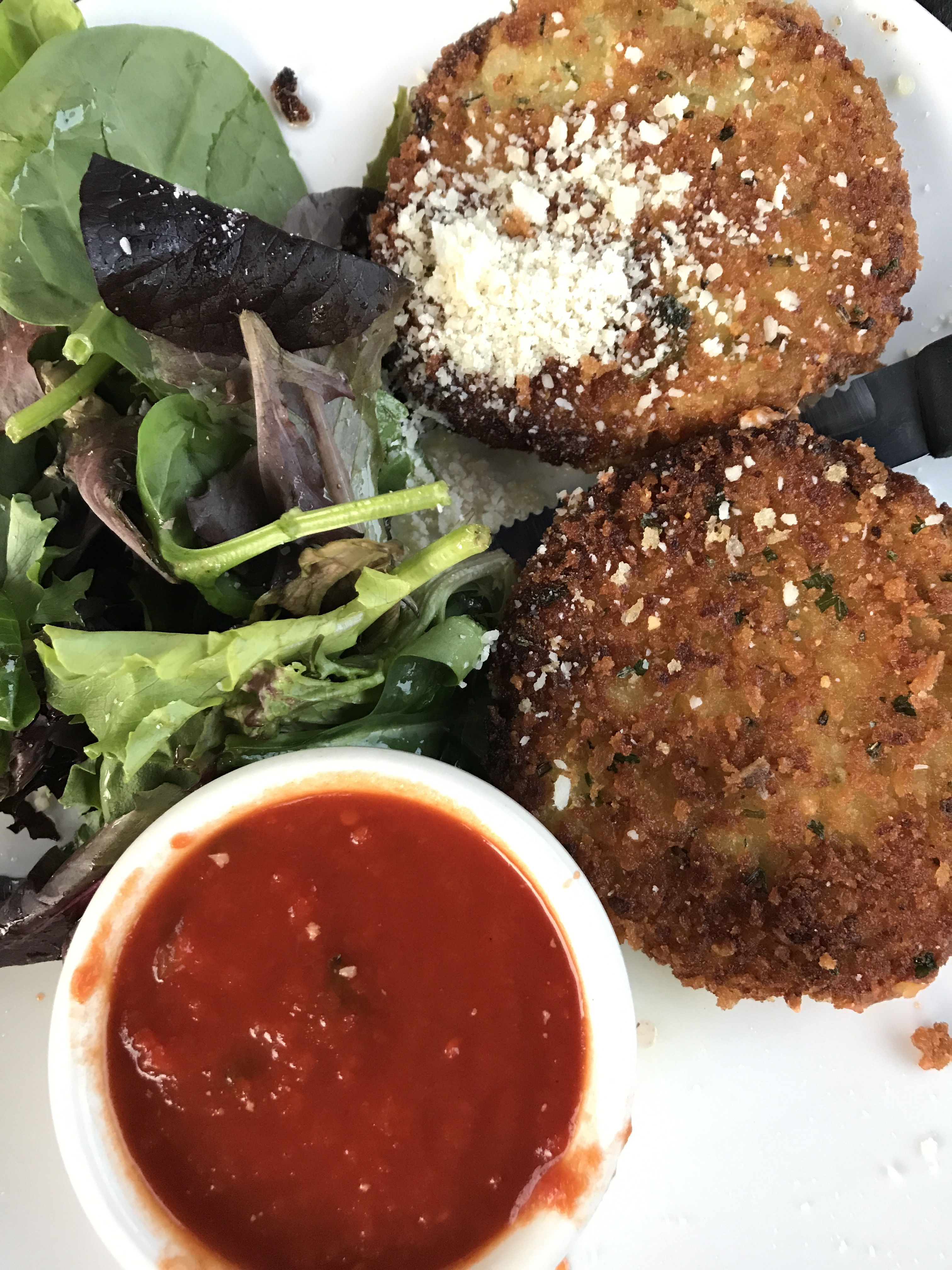 Fried zucchini cakes on a plate with a cup of marinara sauce and greens.