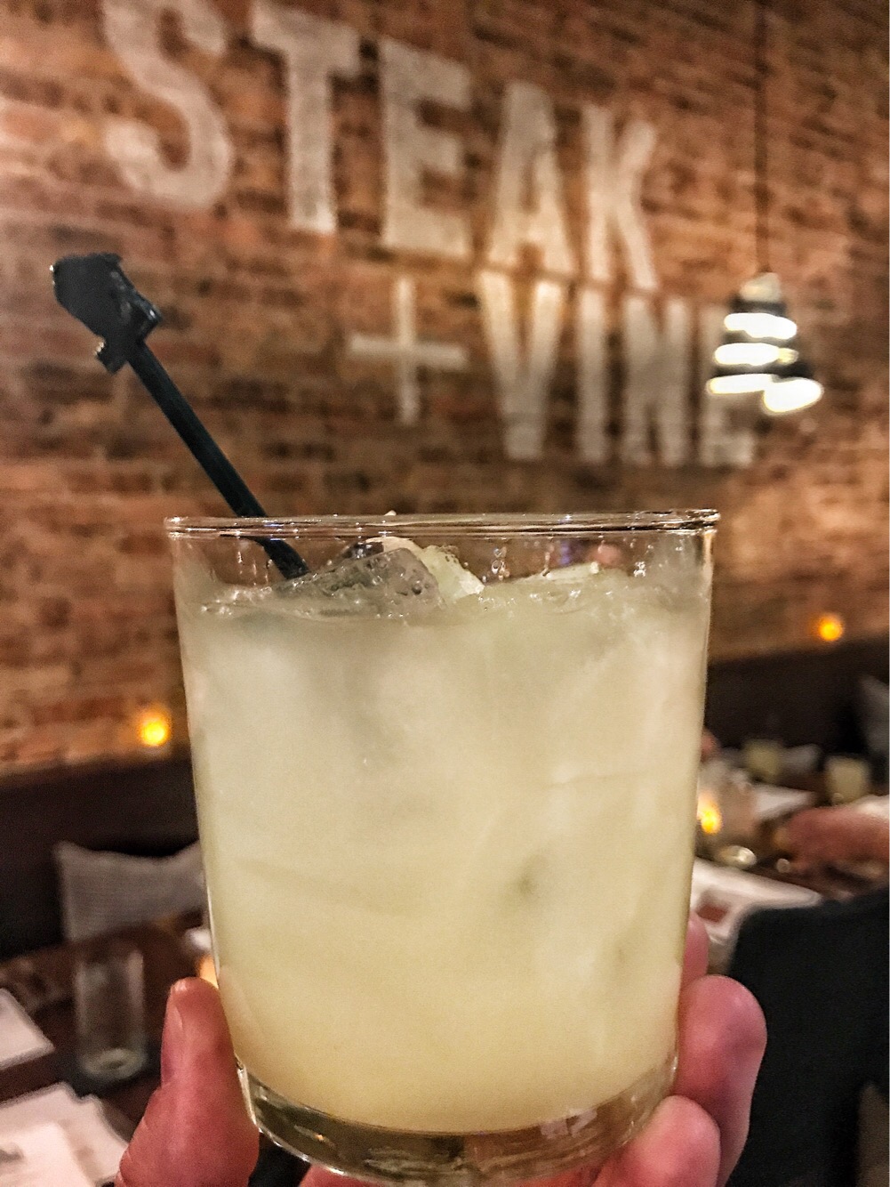 A white cocktail in a glass held in front of a brick wall with Steak + Vine written in big, white block letters.