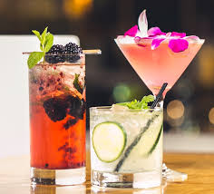Three cocktails. The one on the left is red with blackberries in a tall, highball glass. In the middle is a clear cocktail with lime in an old fashioned glass, and to the right is a pink cocktail with flowers in a martini glass.