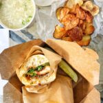 A pork katsua sandwich in a brown cardboard box with a pickle next to potato chips and egg drop soup.