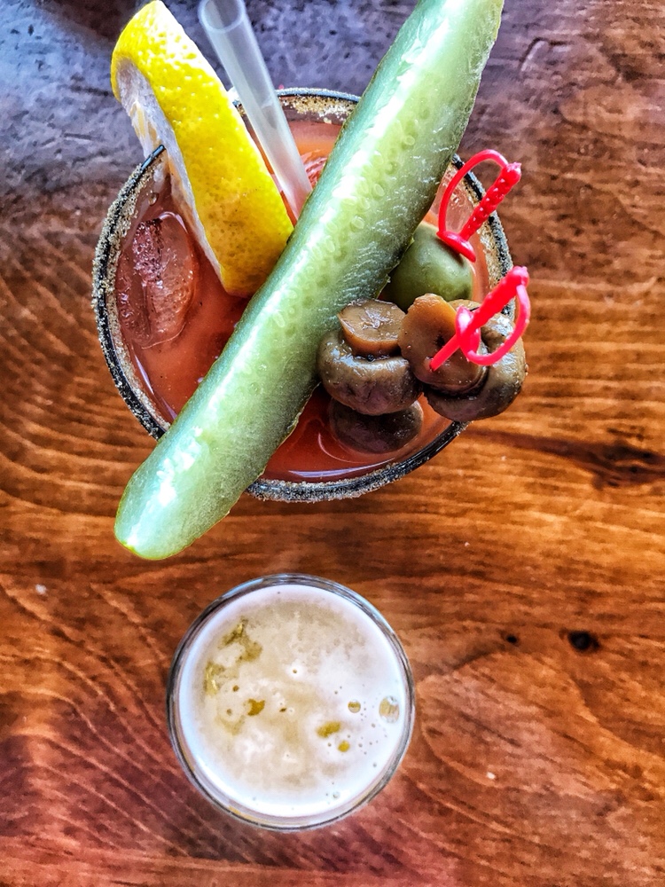 An overhead view of a Bloody Mary with pickles, mushrooms, and lemon next to a smaller beer back.