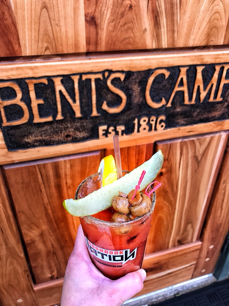 A Bloody Mary being held in front of a wooden sign for Bent's Camp that says "Est 1896" below the name.