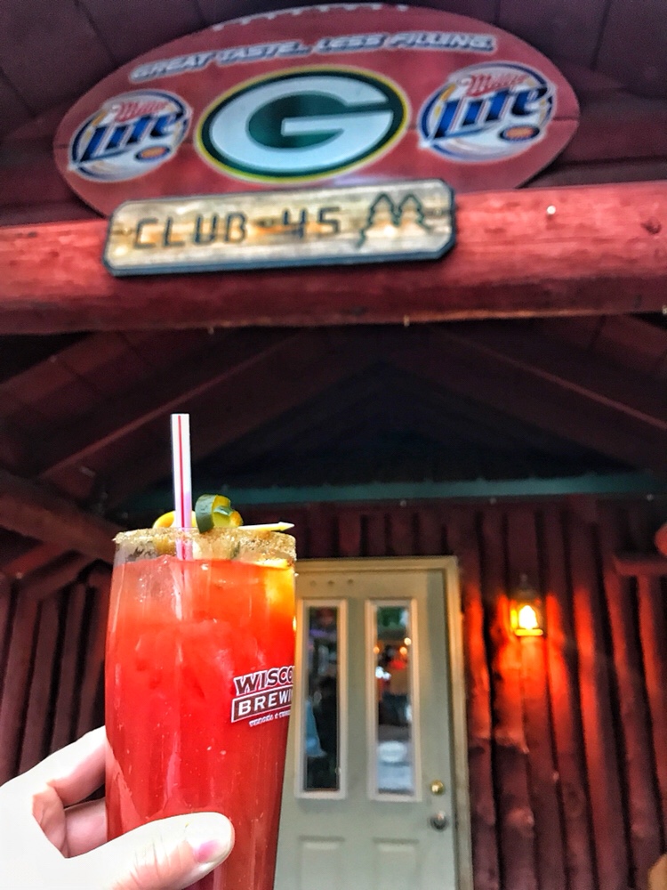 A Bloody Mary held in front of a lodge with a sign that reads "Club 45 M" under a football with a Green Bay Packers logo with Miller Lite logos on either side.