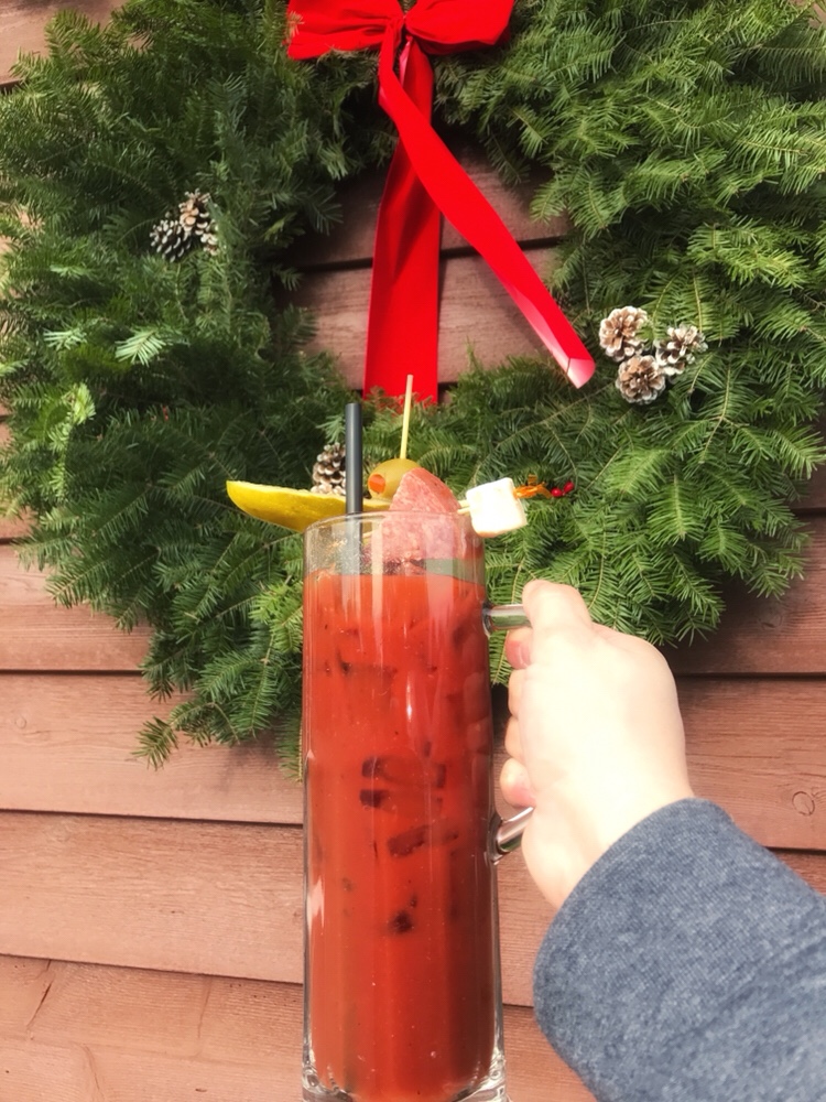 A Bloody Mary topped with pickle, olive, cheese, and salami held in front of a Christmas wreath. 