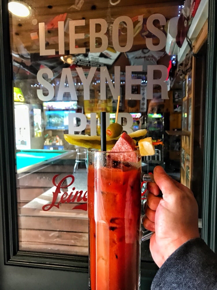 A Bloody Mary in a pint glass topped with a pickle, an olive, a cube of cheese, and a chunk of salami. It is held aloft in front of a window with the words "Liebo's Sayner Pub" on it.
