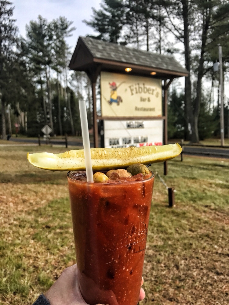 A Bloody Mary topped with a pickle and olives held in front of the sign for Fibber's Bar and Restaurant.