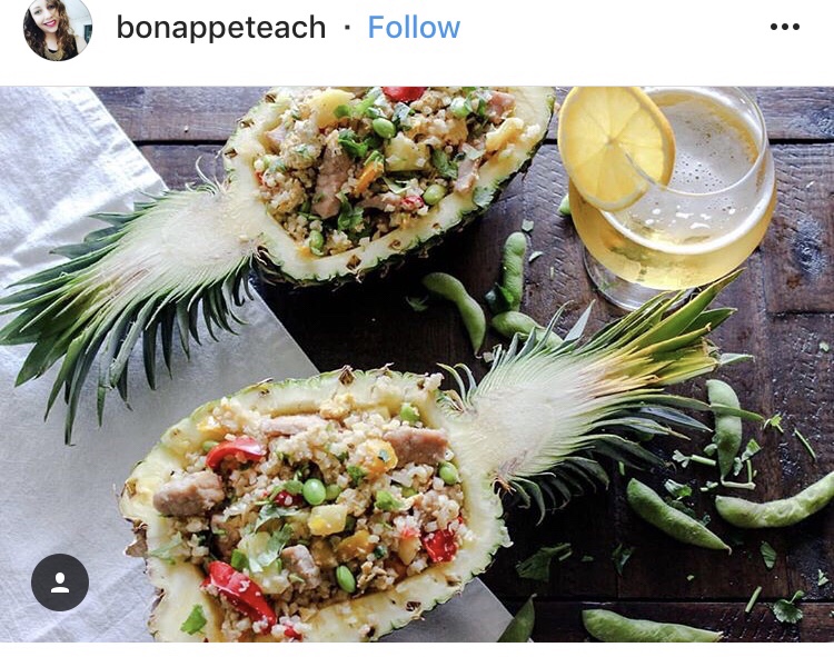 In instagram post from bonappeteach. It features a pineapple sliced in half and each half is filled with cauliflower fried rice. There is a drink with a lemon wedge next to the pineapples.