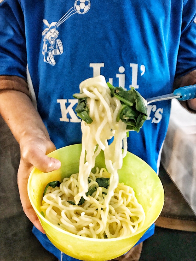 A bowl of ramen noodles and vegetables being held by a child wearing a "Lil' Kicker's" jersey.