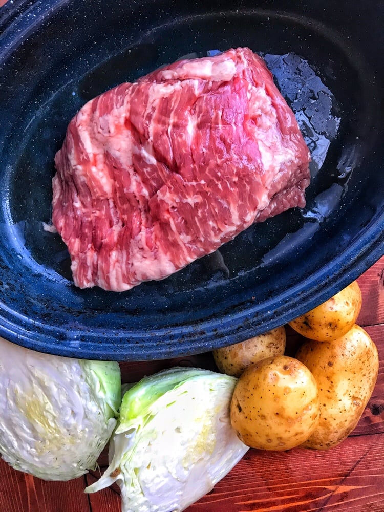 A slab of corned beef in a slow cooking dish with potatoes and cabbage to the side.