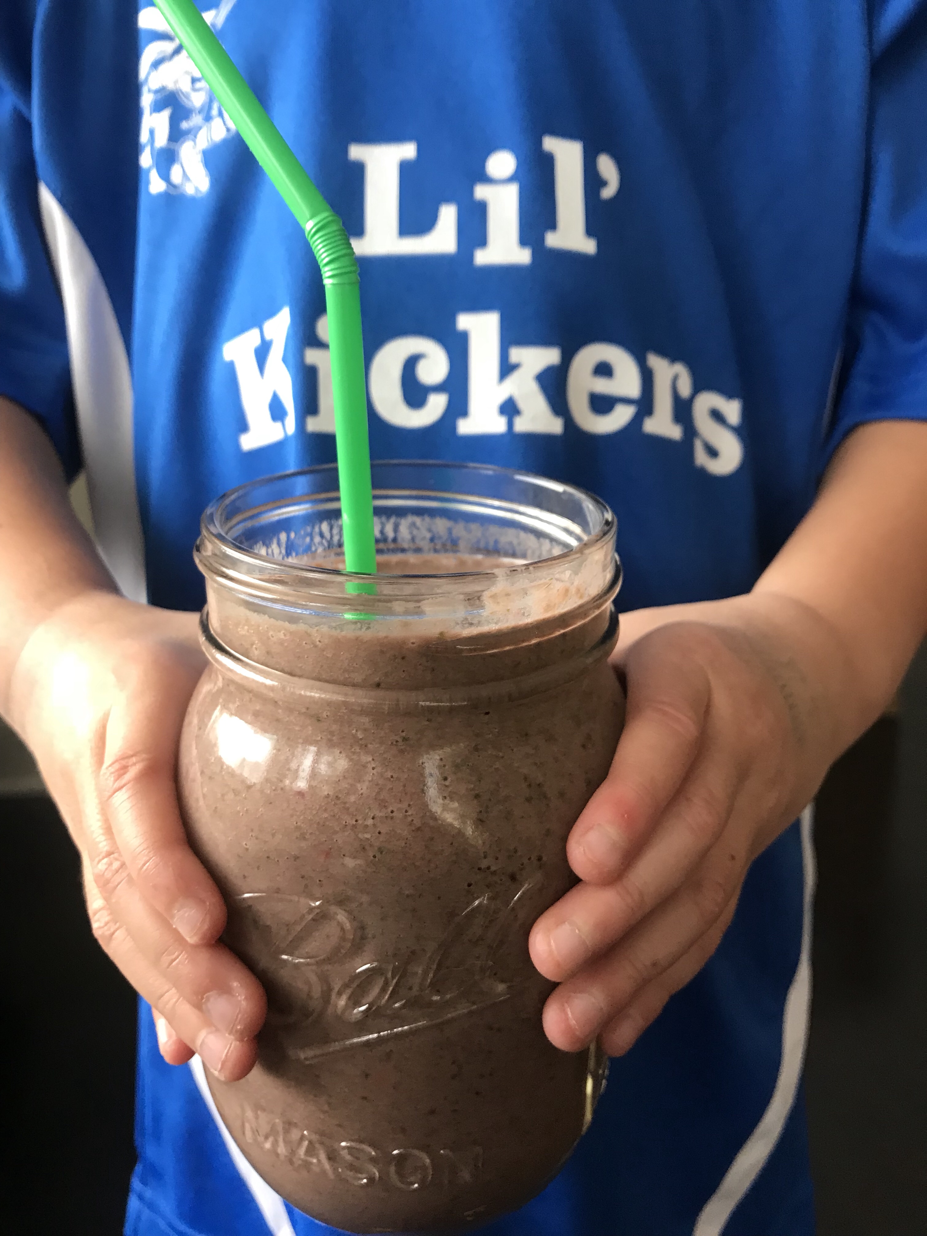 A purpleish smoothie in a mason jar with a green straw being held be a child wearing a "Lil' Kickers" shirt.