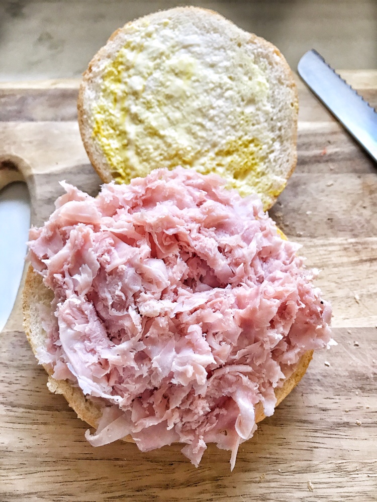 A sliced roll with spread mayo and mustard on one half and shaved deli ham on the other.