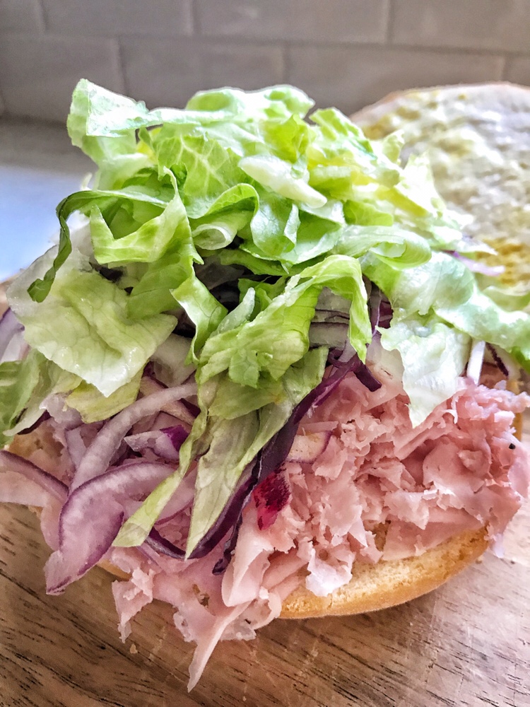 A sliced roll with mayo and mustard spread on one half and shaved ham, red onion, and shredded lettuce on the other half.