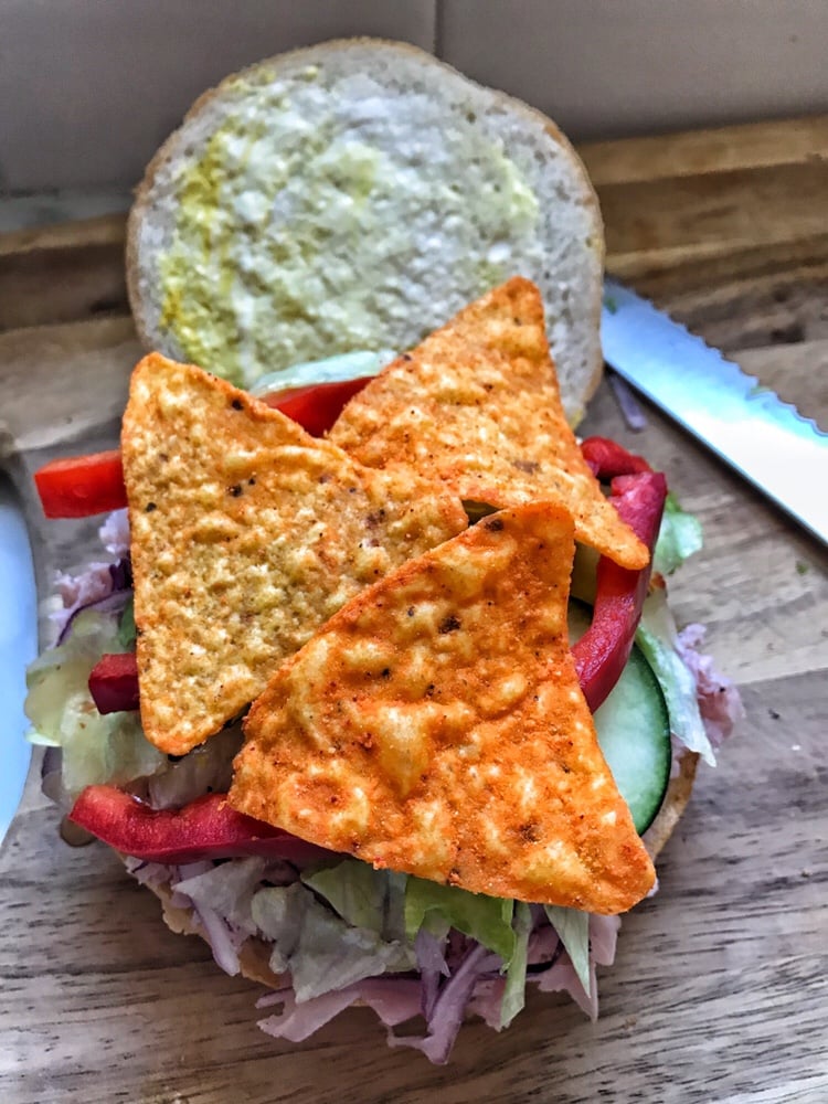 A sliced roll with mayo and mustard spread on one half and shaved ham, red onion, shredded lettuce, sliced red pepper, and Doritos on the other half.