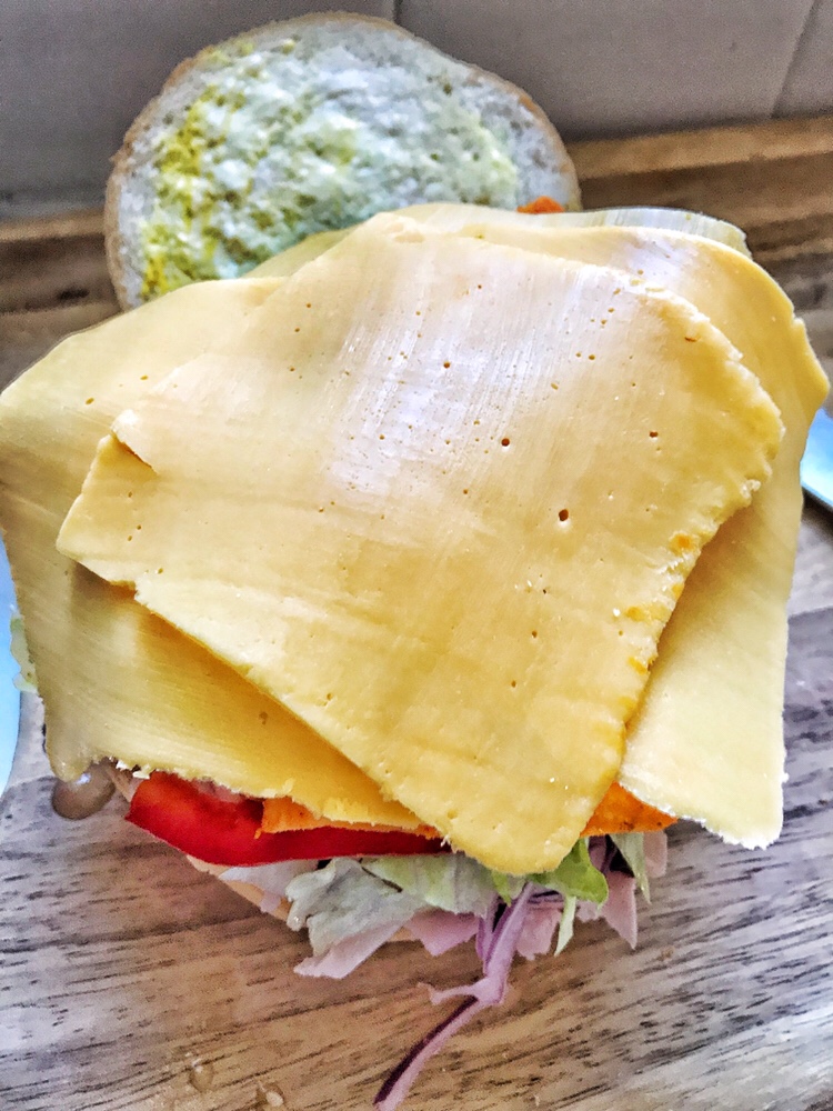 A sliced roll with mayo and mustard spread on one half and shaved ham, red onion, shredded lettuce, sliced red pepper, Doritos, and sliced cheese on the other half.