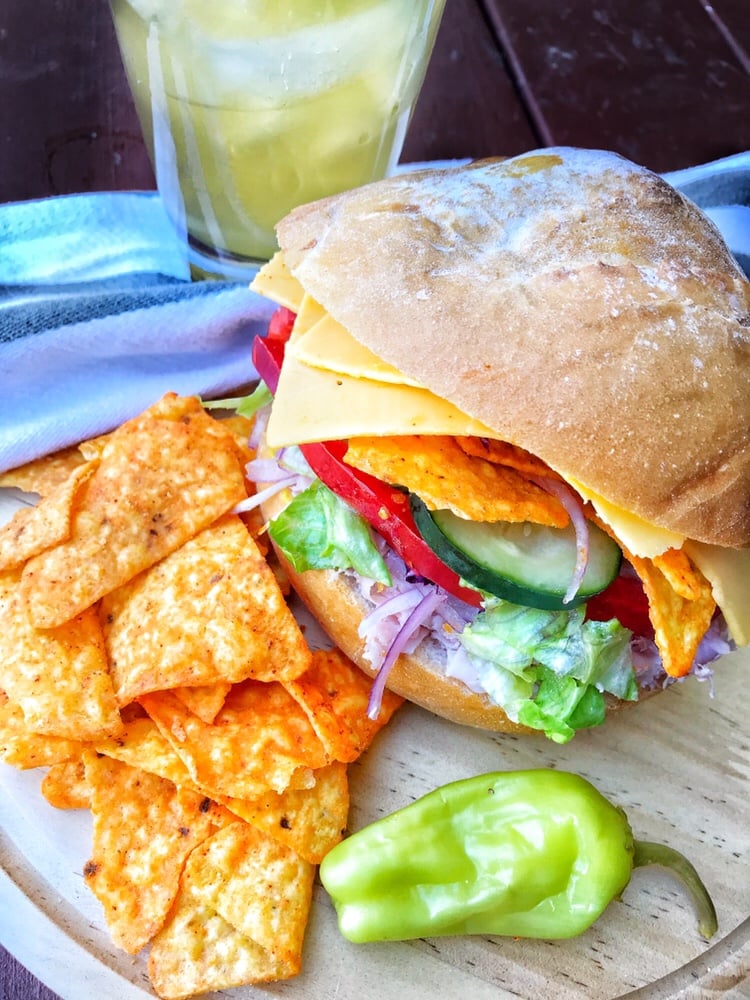 A ham sandwich with veggies, cheese, and Dorito chips is on a circular wooden cutting board with more Doritos and a pepperoncini pepper next to it.