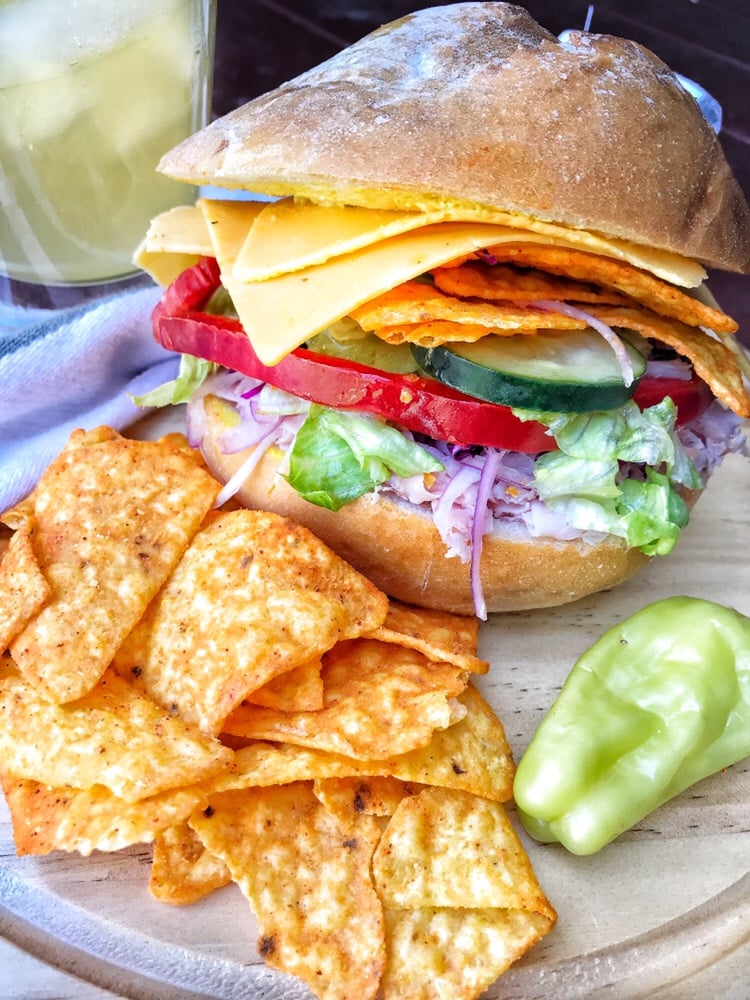 A ham sandwich with veggies, cheese, and Dorito chips is on a circular wooden cutting board with more Doritos and a pepperoncini pepper next to it.