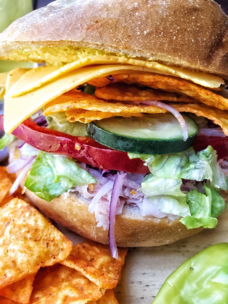 A close up view of a ham sandwich with lettuce, red onion, cucumbers, red peppers, Doritos, cheese, and mustard.