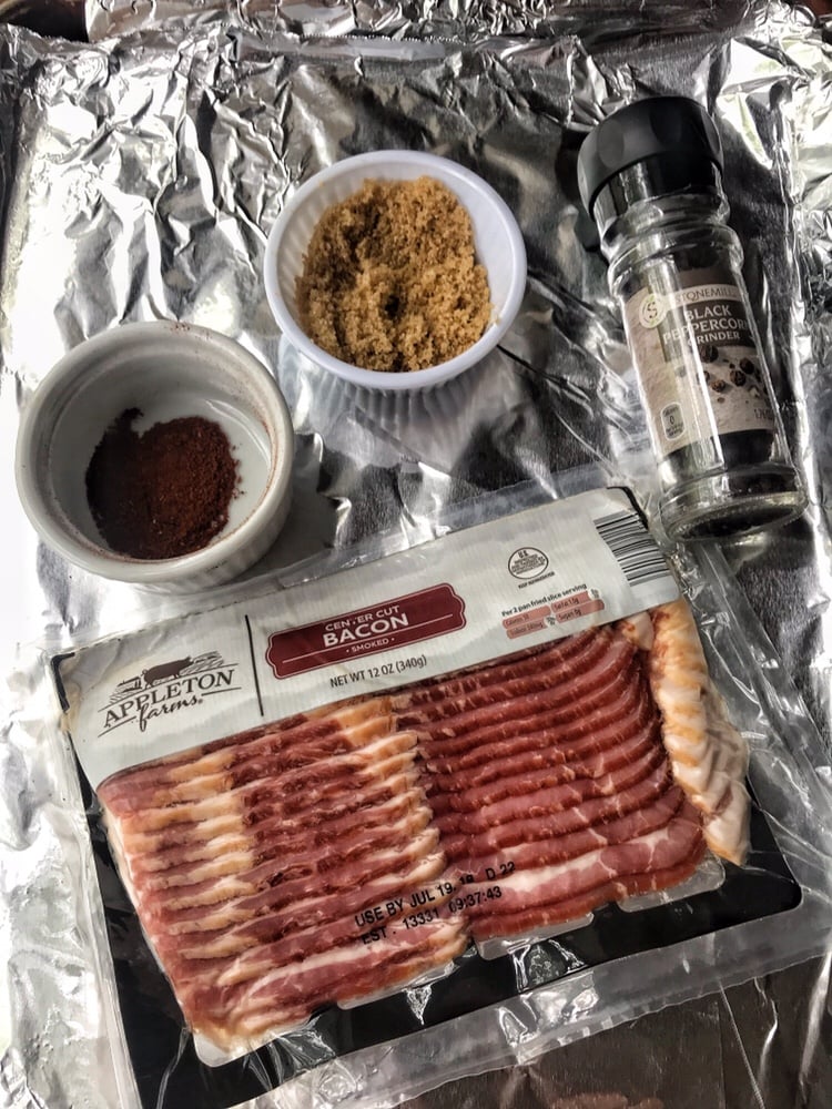 A baking sheet covered in foil. On top is a package of raw bacon, a black pepper grinder, a cup of brown sugar, and a cup of chili powder.