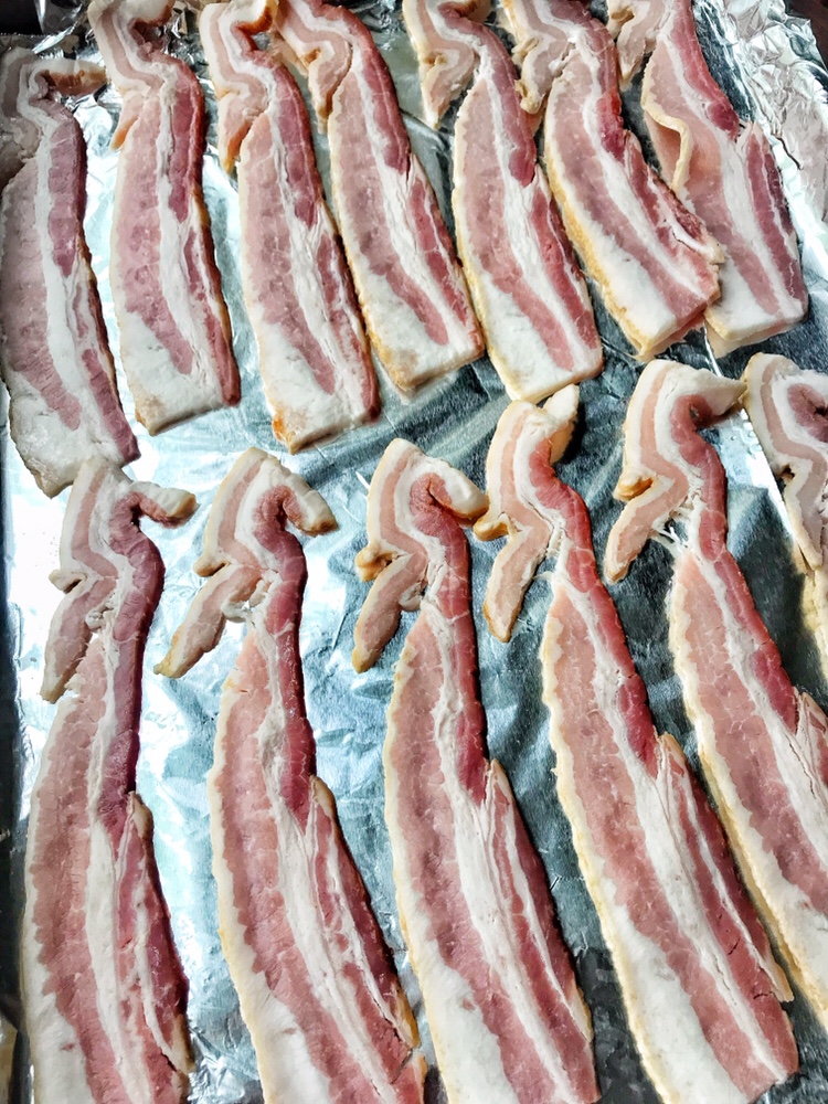 Pieces of raw bacon spread out on a foil-covered baking sheet.