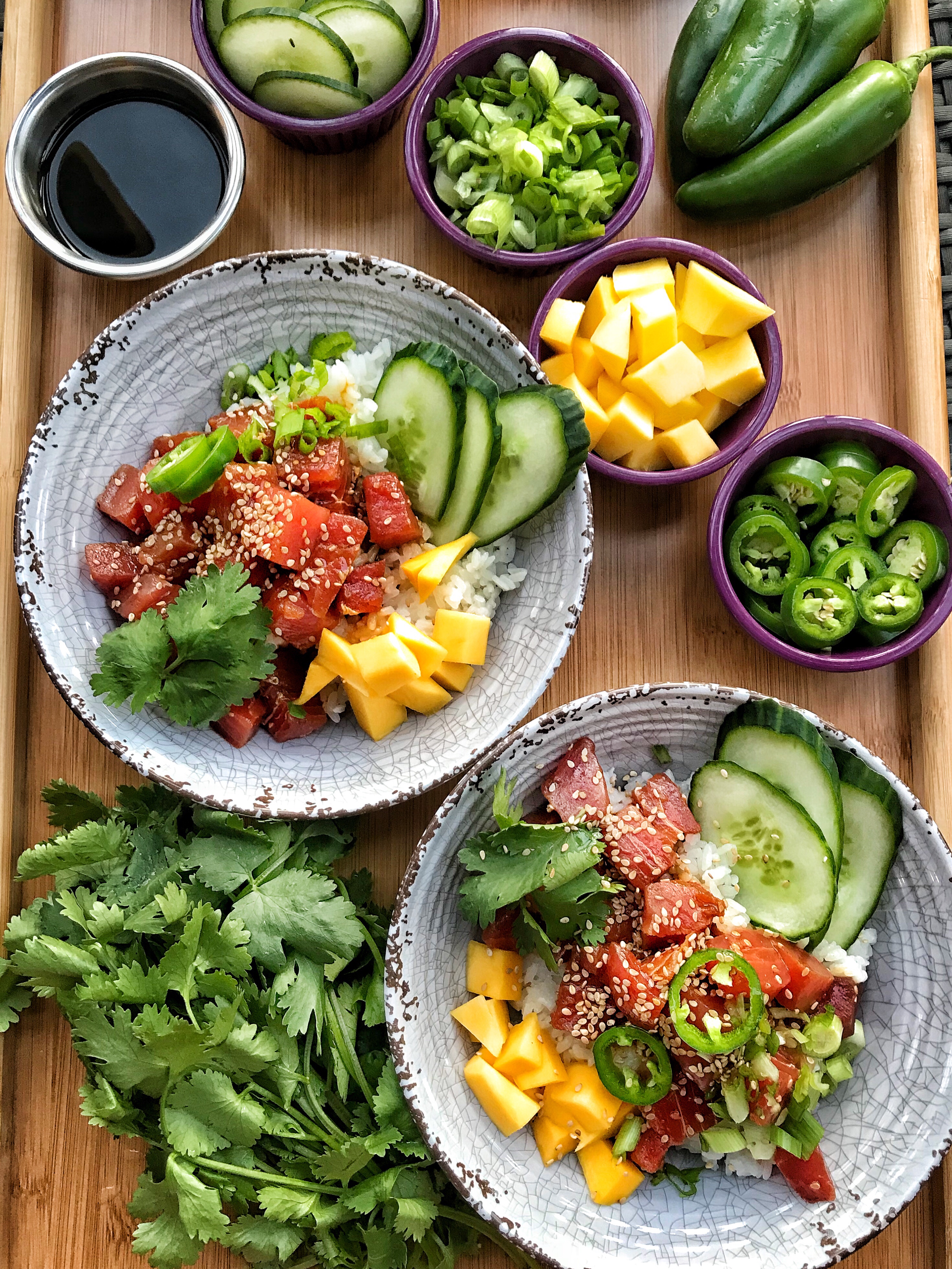 Two rice bowls with cubes of tuna, mango, cucumber slices, and sesame seeds on a wooden cutting board along with soy sauce and jalapenos.