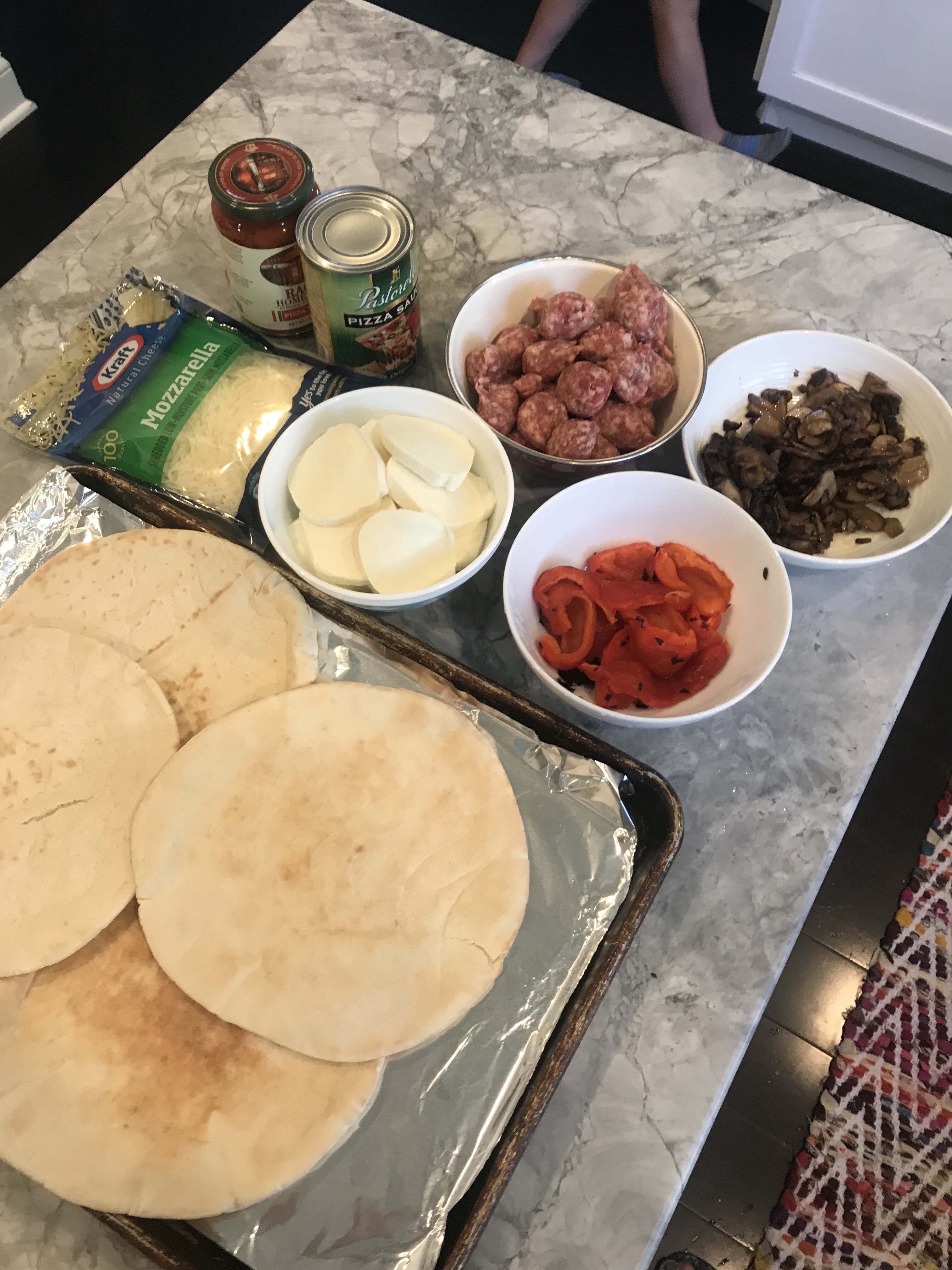 The ingredients for the pizza including pita bread, mozzarella, shredded mozarella, peppers, sausage, mushrooms, and tomato sauce