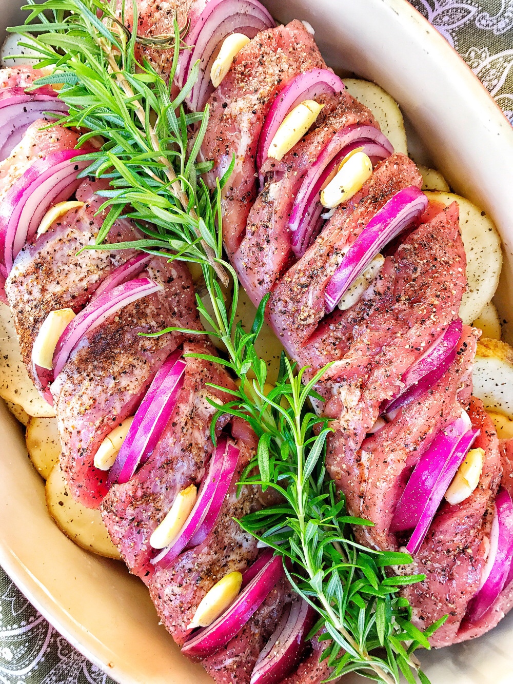 Raw pork tenderloins with slits cut into them. Onion slices and garlic pieces are inserted into the slits. There is rosemary in between the two tenderloins and potato slices underneath them.