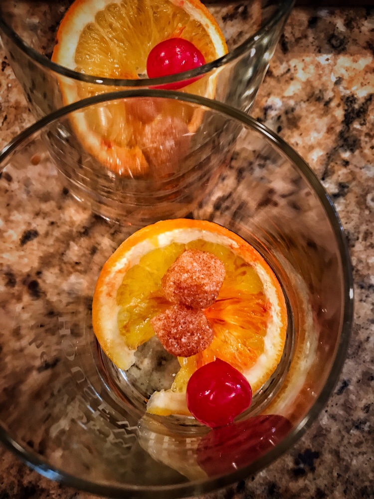 Two glasses with and orange slice, cherry, and couple sugar cubes each inside them.