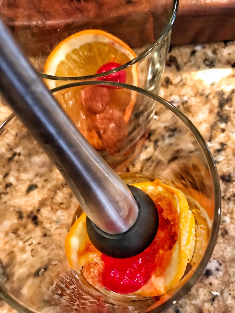 A muddler being used to mash up cherry, orange, and sugar cubes together in a glass.