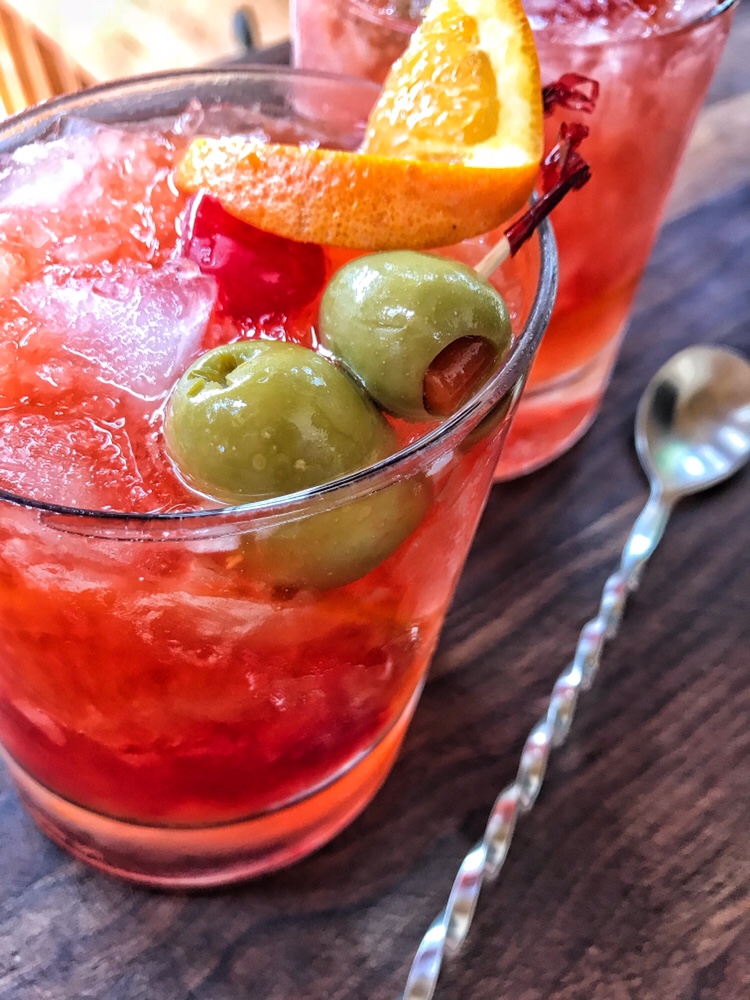 A close up of a brandy old fashioned. It is garnished with a cherry, and orange twist, and two olives on a toothpick.