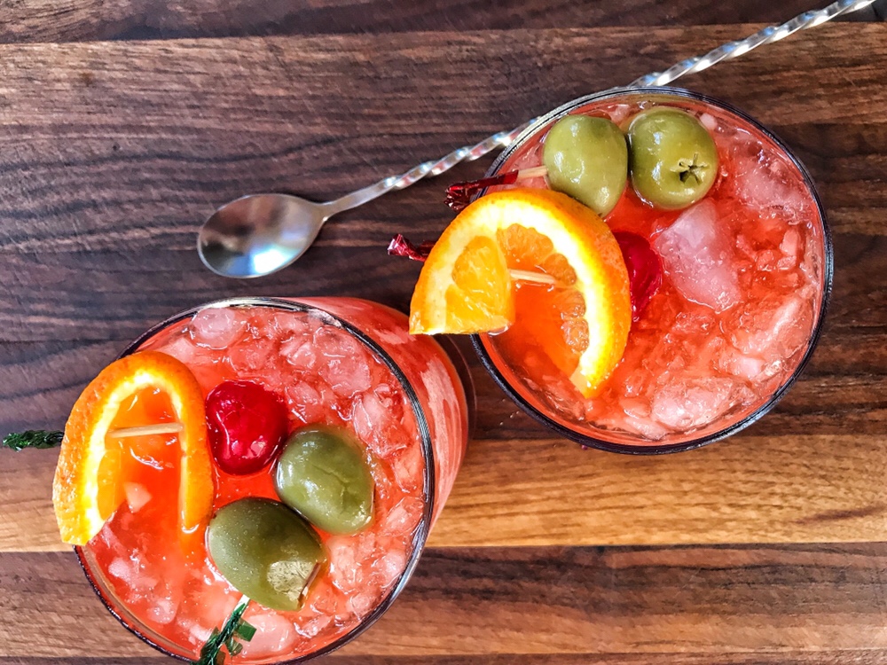 Two brandy old fashioneds with olives, cherries, and an orange slice on top seen from above.