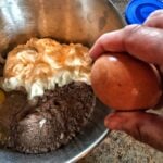 An egg being cracked against the side of a metal bowl which contains cake mix and mayo.
