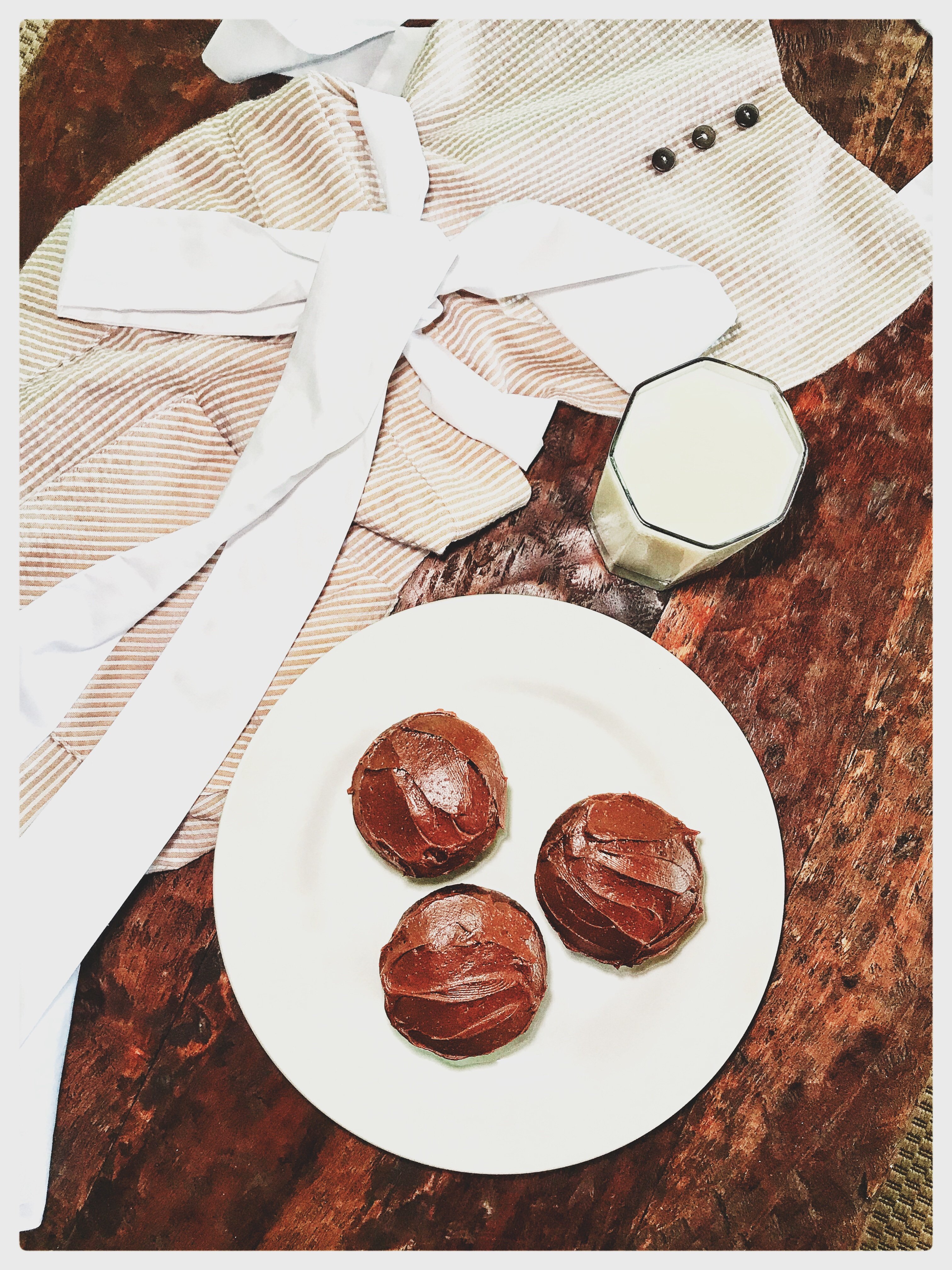 An apron next to a glass of milk and a white plate with three chocolate cupcakes.
