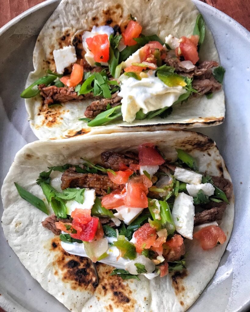 Two steak tacos with tomato, cheese, onion cilantro, and sour cream.