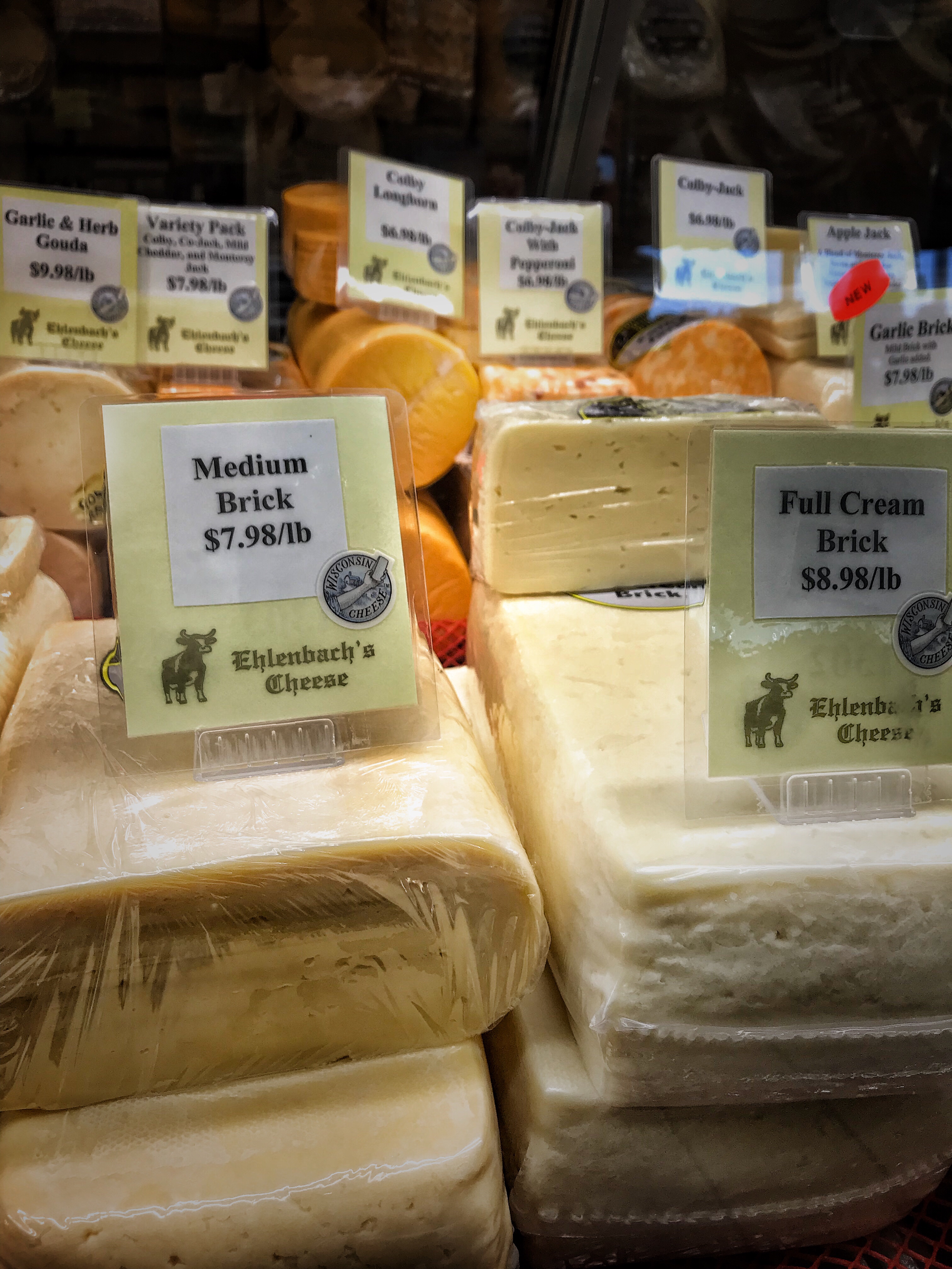 A collection of cheeses for sale. In the foreground is a medium brick for 7.98 a pound and a full cream brick for 8.98 a pound.
