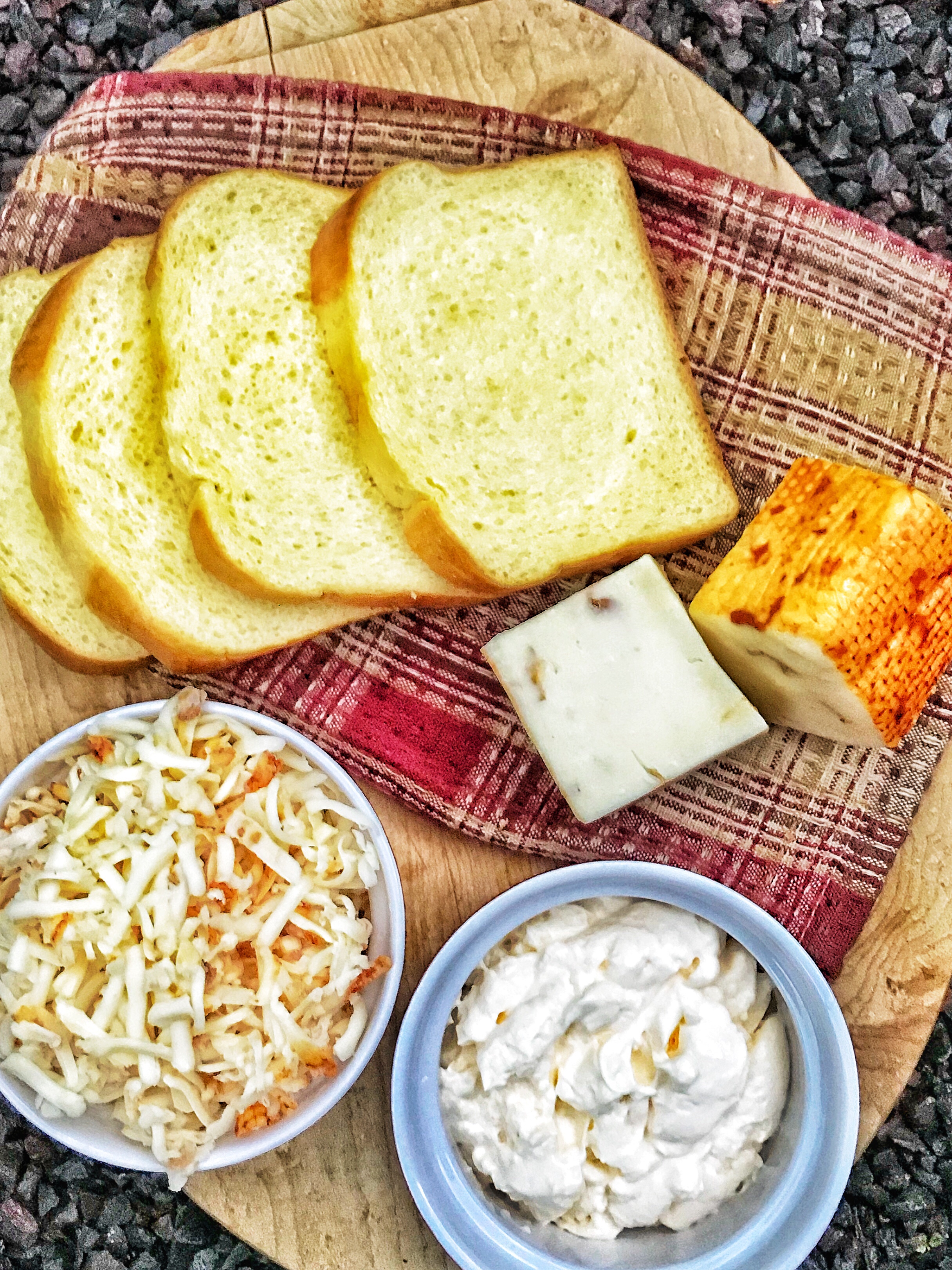 Slices of brioche bread on a cutting board next to a block of cheese, shredded cheese, and mayonnaise in a blue bowl.