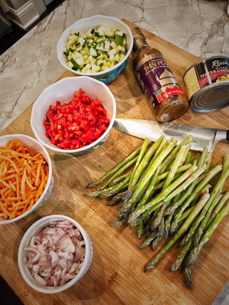 A cutting board with the ingredients for Thai lettuce wraps, including asparagus, chopped onion, carrot, zucchini, and diced red pepper next to a can of water chestnuts and Thai peanut sauce.