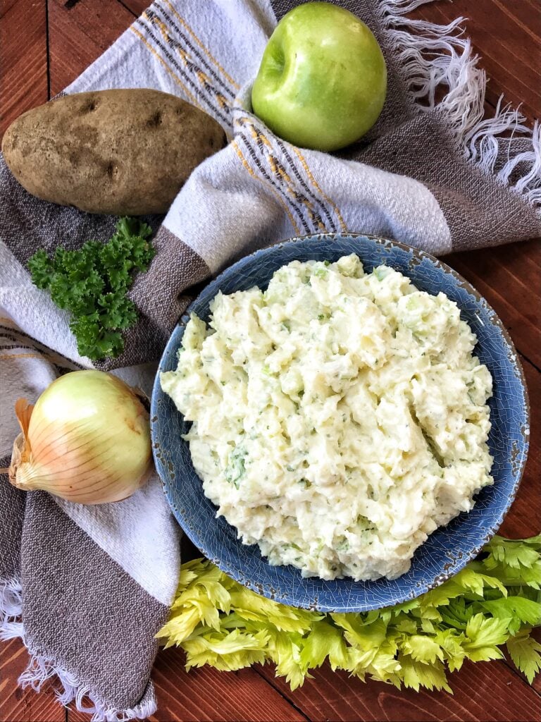 A blue bowl of Spring Potato Salad next to vegetables and a hand towel.