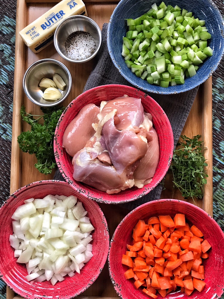 Raw chicken in a red bowl surrounded by the other ingredients for the dish, including chopped onion, celery, carrots, garlic, salt, pepper, and a stick of butter.