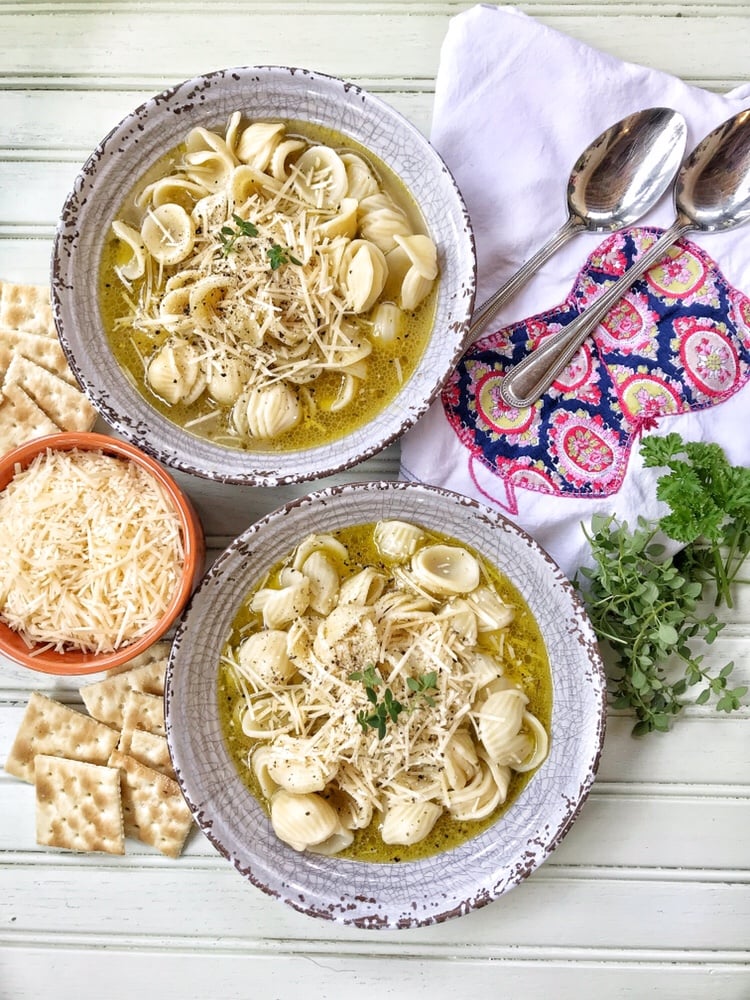 Two bowls of buttered herb noodles in chicken broth alongside spoons, crackers, and herbs.