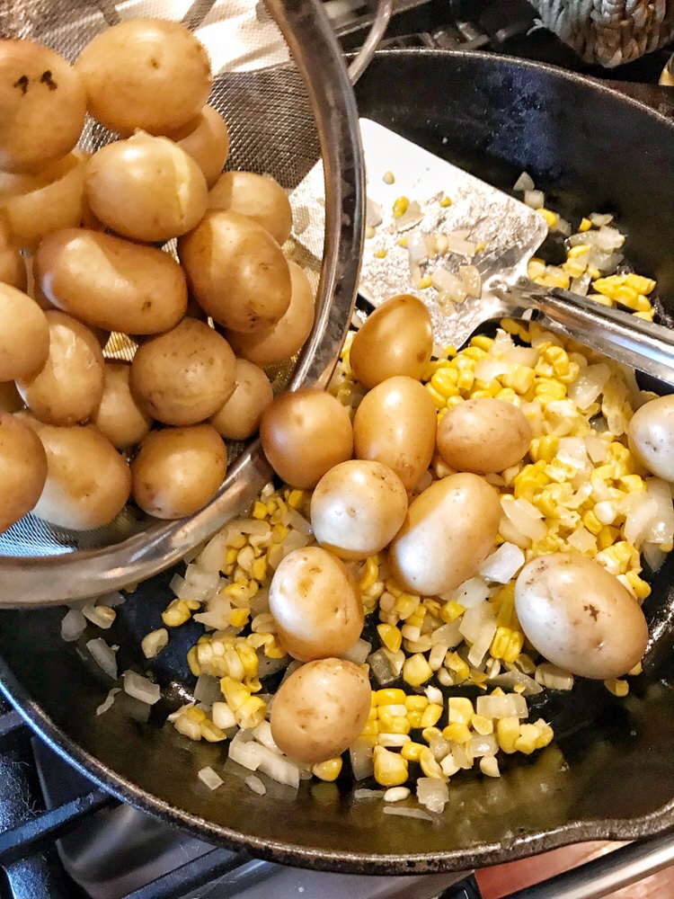 Potatoes being poured into a skillet with corn and onions.