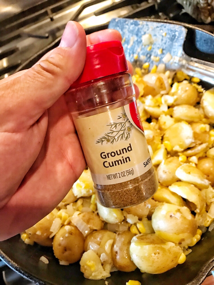 Ground cumin held over the potatoes, corn, and onions in a pan.