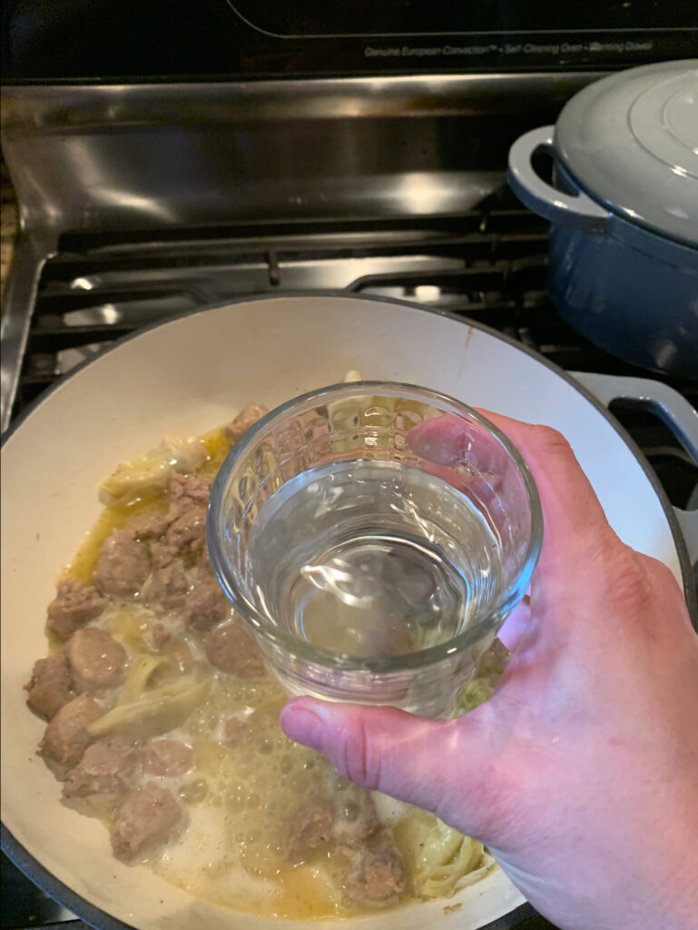 A cup of water about to be poured into artichoke and meats cooking in a pan