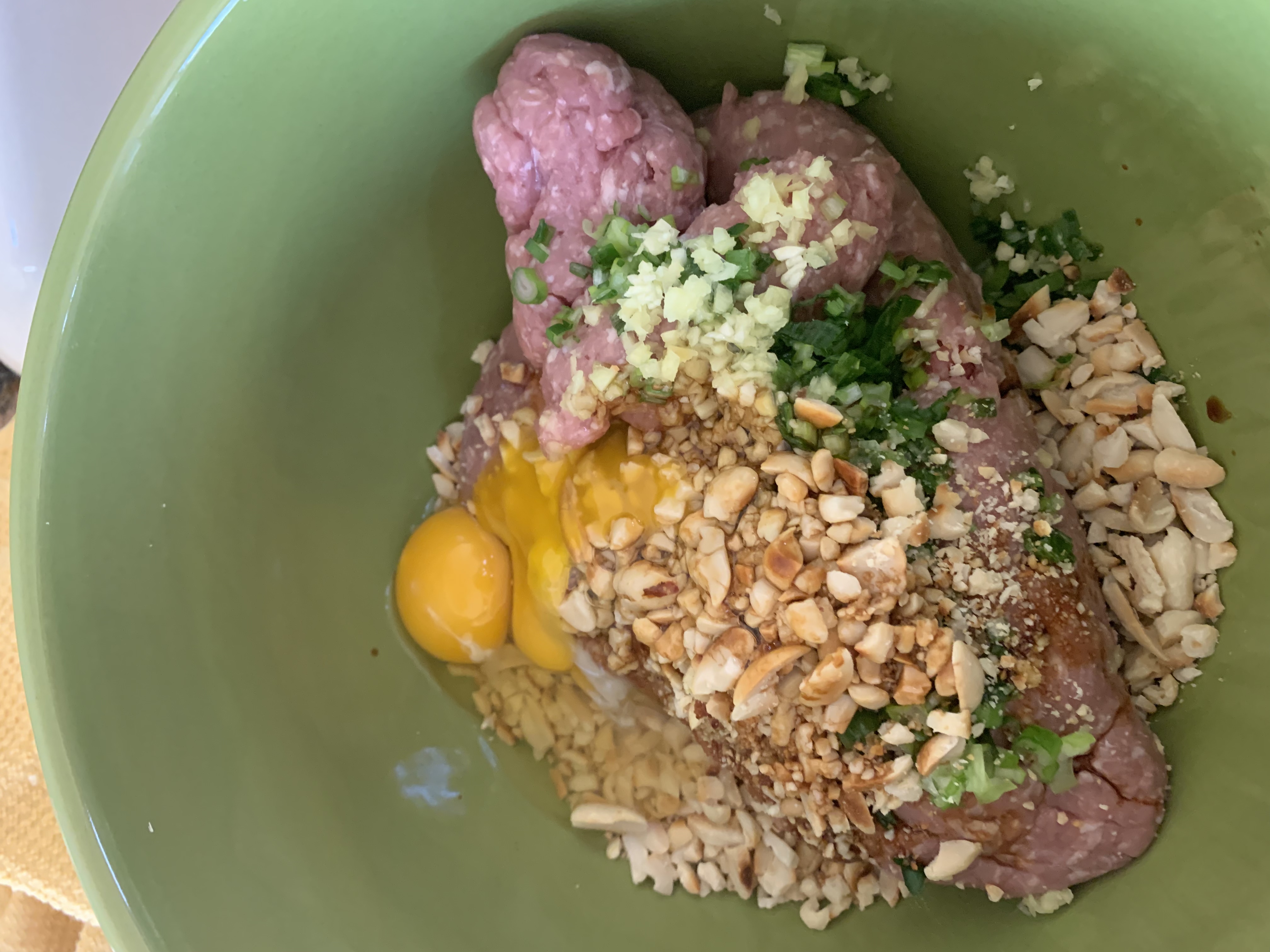 Raw ground pork, eggs, peanuts, green onions and ginger in a green bowl with garlic, waiting to be mixed to make meatballs.