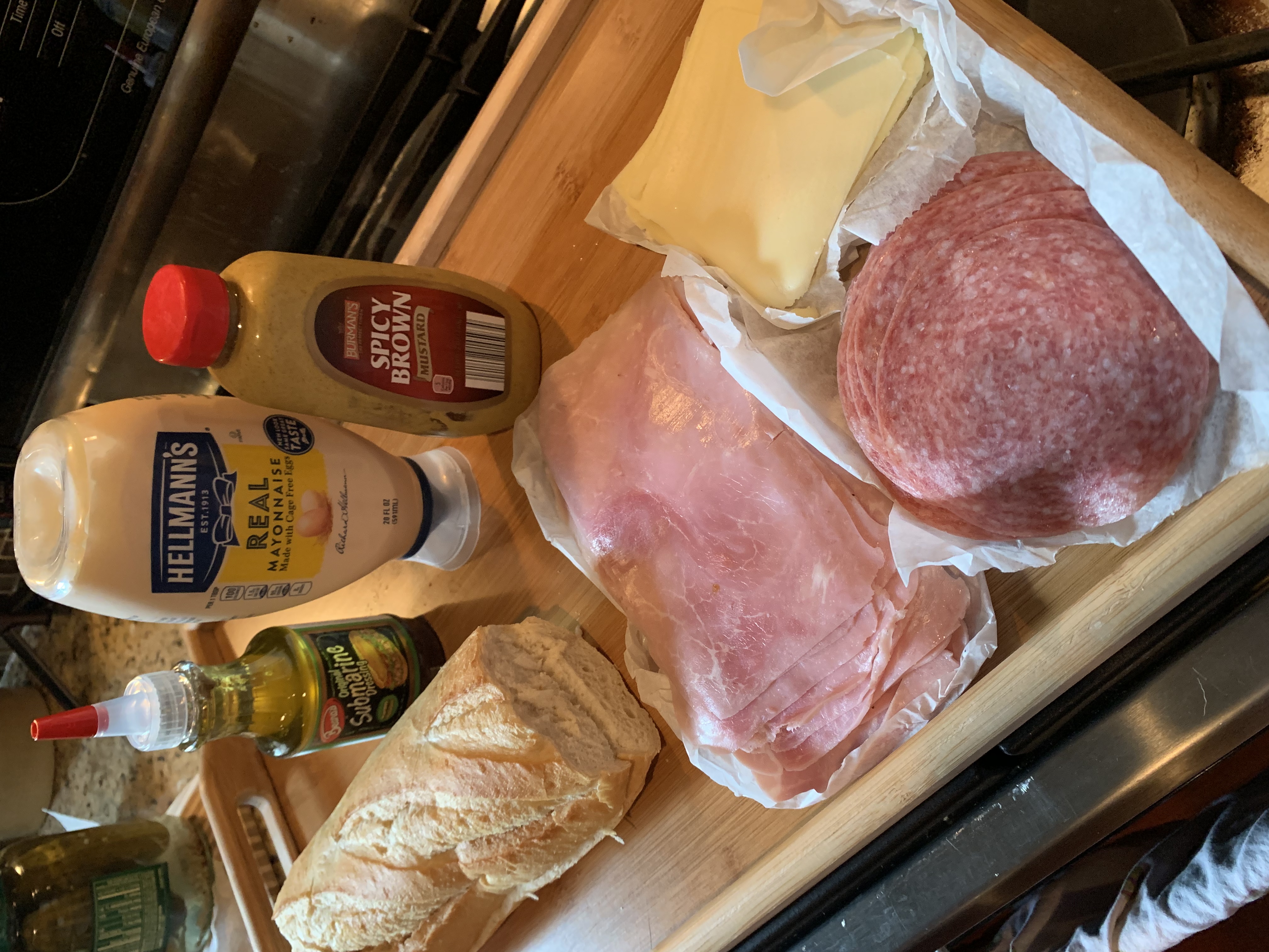 A photo of sandwich ingredients including French bread, Beano's sandwich dressing, mayo, spicy brown mustard, sliced ham, sliced salami, and sliced cheese