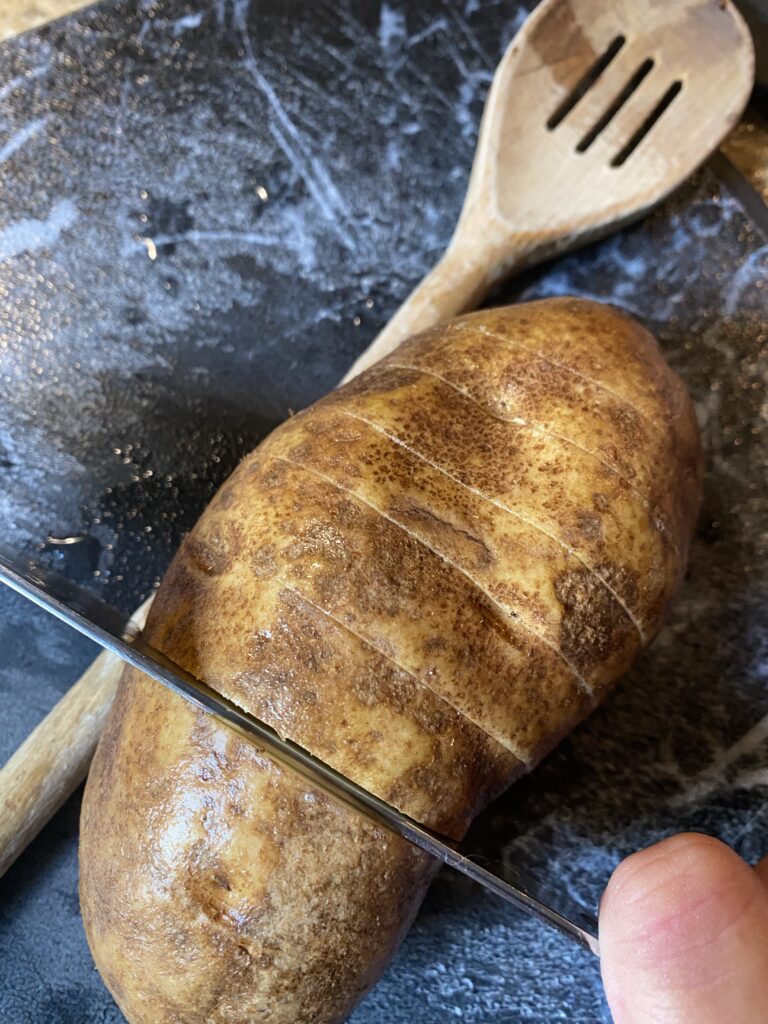 A potato shown next to the handle of a wooden spoon and a knife so that the knife doesn't cut all the way through the potato.