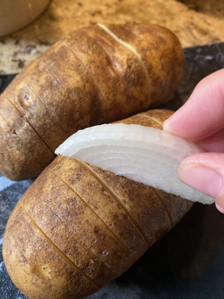 A slice of onion held in front of potatoes with slices cut partway through them