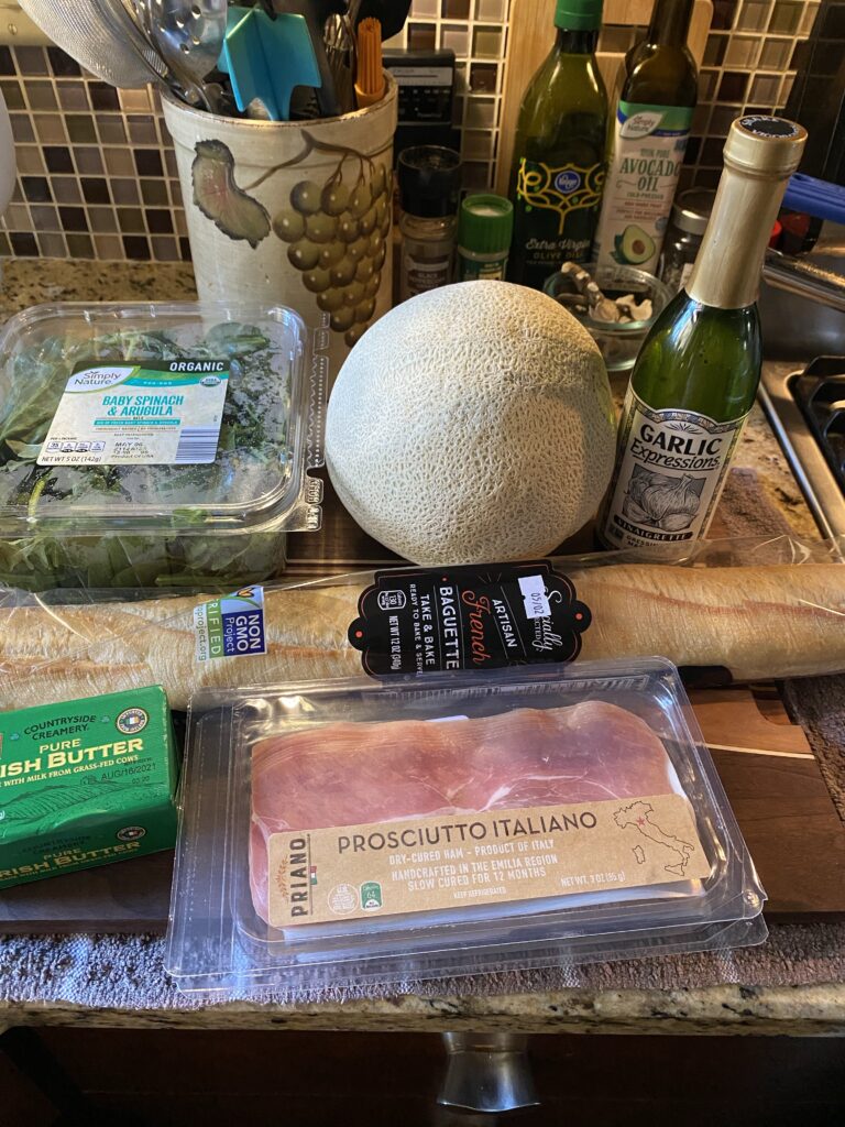 The ingredients for the sandwich, including cantaloupe, baguette, butter, prosciutto, Garlic Expressions dressing, and baby spinach and arugala